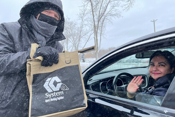 A masked IC System employee poses handing a goody bag to another IC system employee sitting in her car